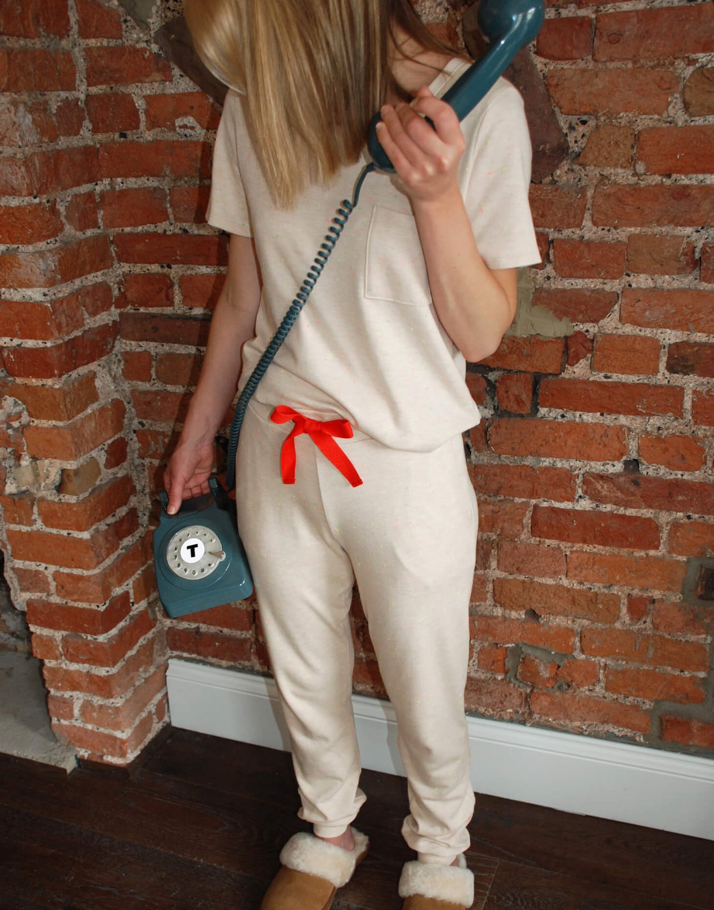 Tessie Clothing Confetti Oat Pocket T-Shirt and matching Confetti Oat Pyjama Trousers Set | Model stood in front of a brick wall wearing Tessie Clothing's Confetti Oat Pocket T-Shirt Pyjama Set while wearing chestnut brown sheepskin slipper mules. Model is using a retro blue telephone.