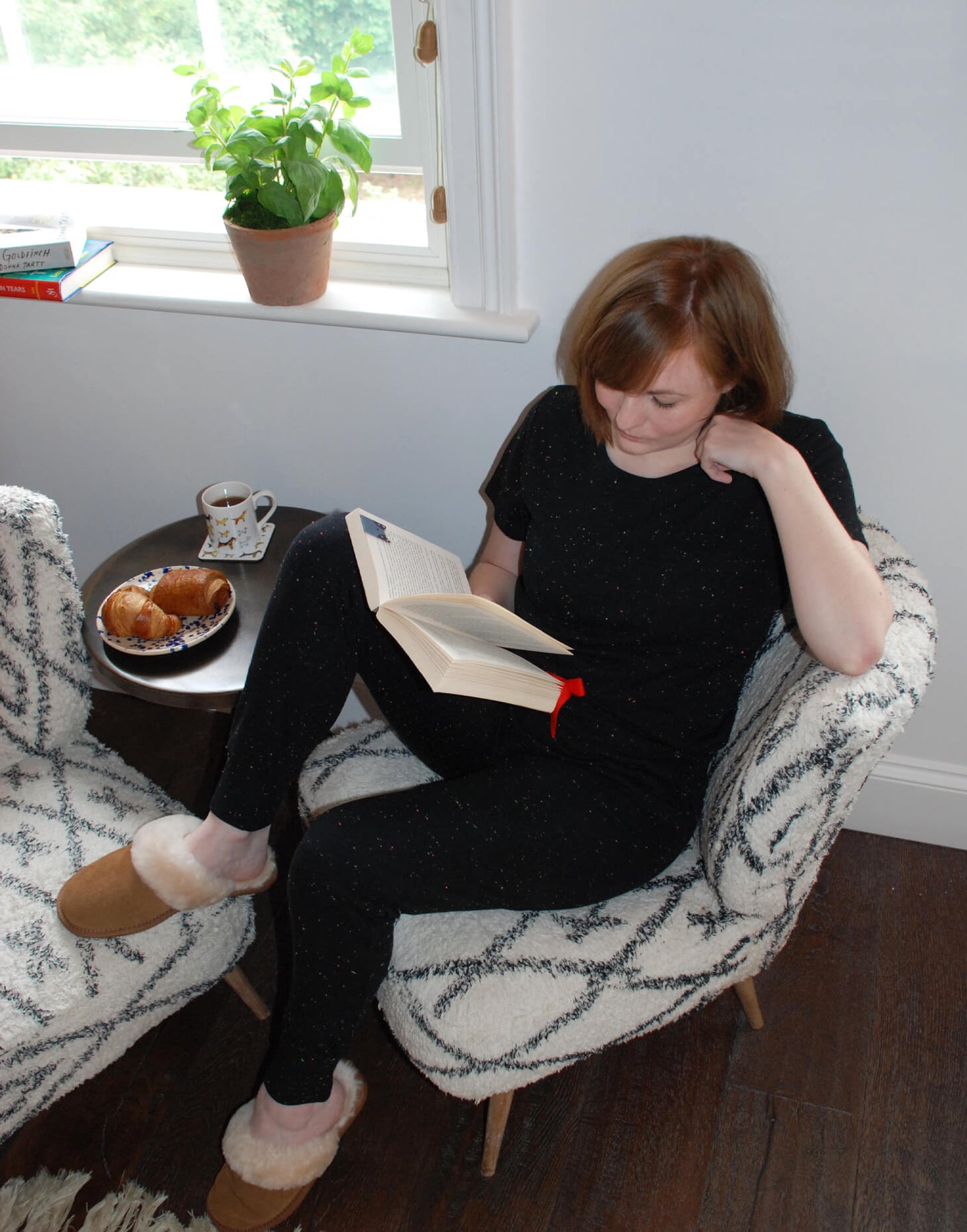 Tessie Clothing Confetti Black Pocket T-Shirt and matching Confetti Black Pyjama Trousers Set | Model sat on white and grey chair wearing Tessie Clothing's Confetti Black Pocket T-Shirt Pyjama Set with some chestnut brown sheepskin slipper mules on. Model has a side table next to her where she has a dog print mug and coaster next to it and a plate of croissants. Model is reading a book with a Magnetic Dog Bookmark being used.