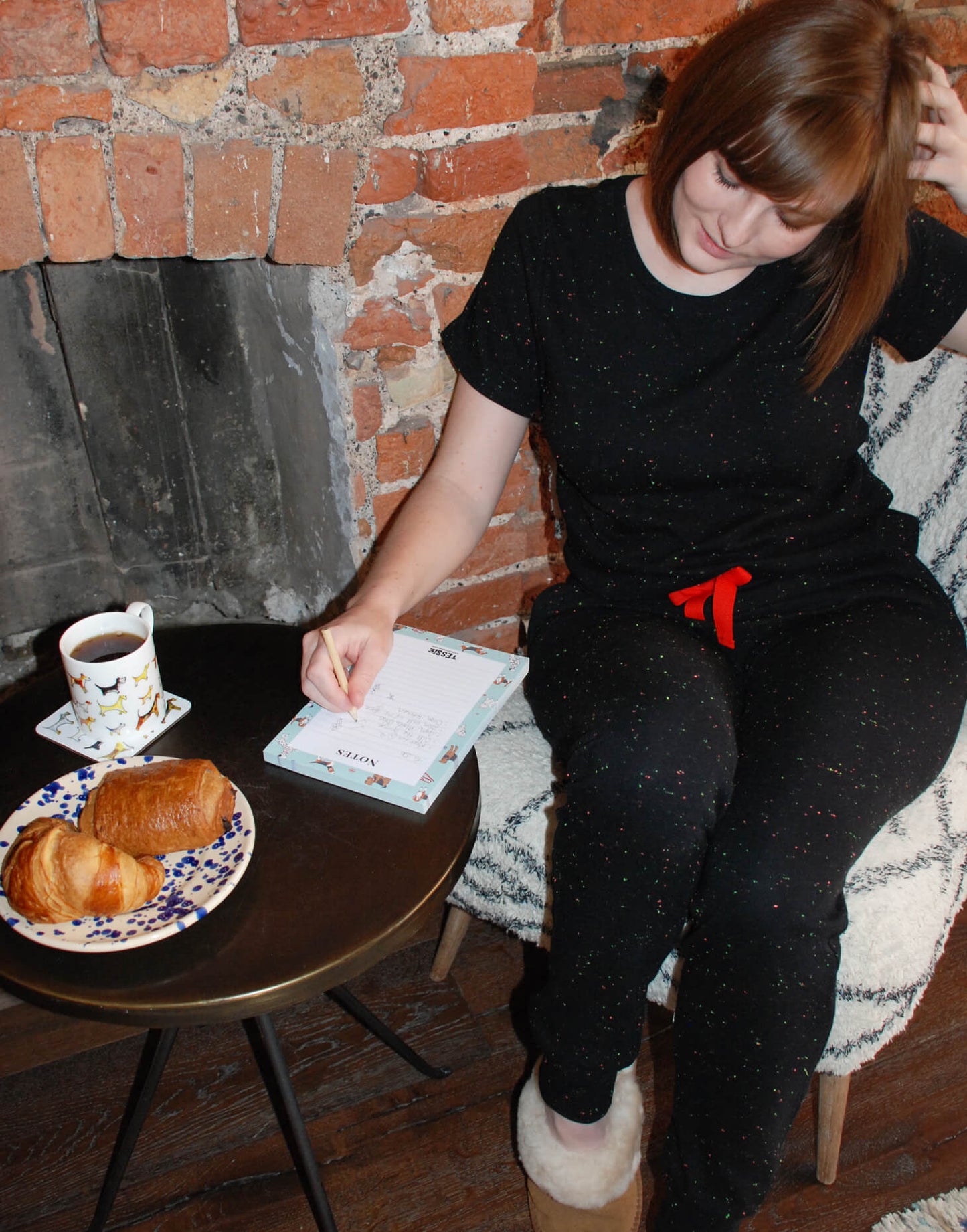 Tessie Clothing Confetti Black Pocket T-Shirt and matching Confetti Black Pyjama Trousers Set | Model sat on white and grey chair wearing Tessie Clothing's Confetti Black Pocket T-Shirt Pyjama Set with some chestnut brown sheepskin slipper mules on. Model has a side table next to her where she is writing on a to do list on an A5 Poppy Dog Print Notepad with a dog print mug and coaster next to it and a plate of croissants.
