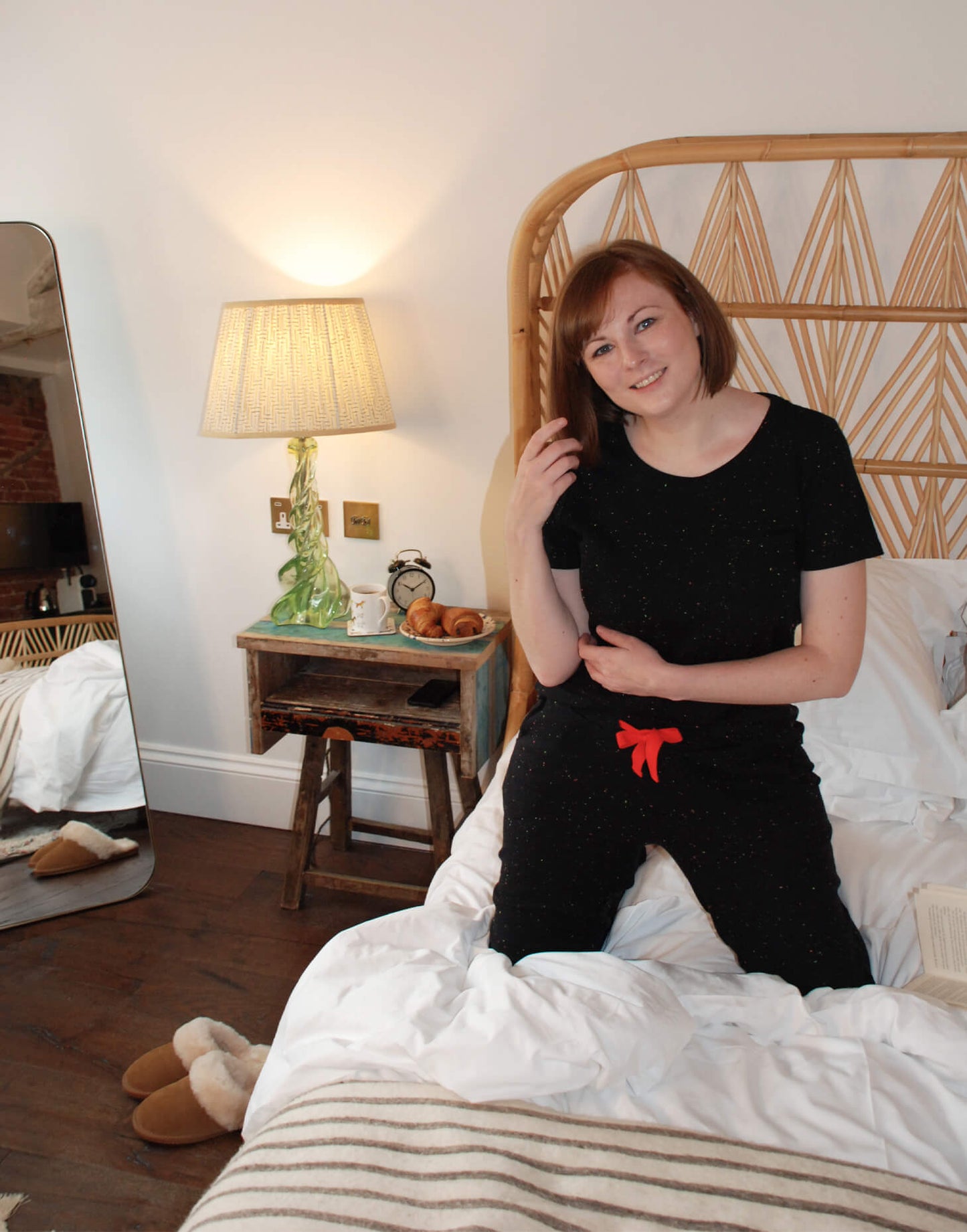 Tessie Clothing Confetti BlackPocket T-Shirt and matching Confetti Black Pyjama Trousers Set | Model knelt on white bedding with a light wood cane headboard wearing Tessie Clothing's Confetti Black Pocket T-Shirt Pyjama Set with some chestnut brown sheepskin slipper mules next to the bed. Next to the bed is a rustic side table with a croissant on it, retro lamp, retro alarm clock and a dog print mug and coaster.