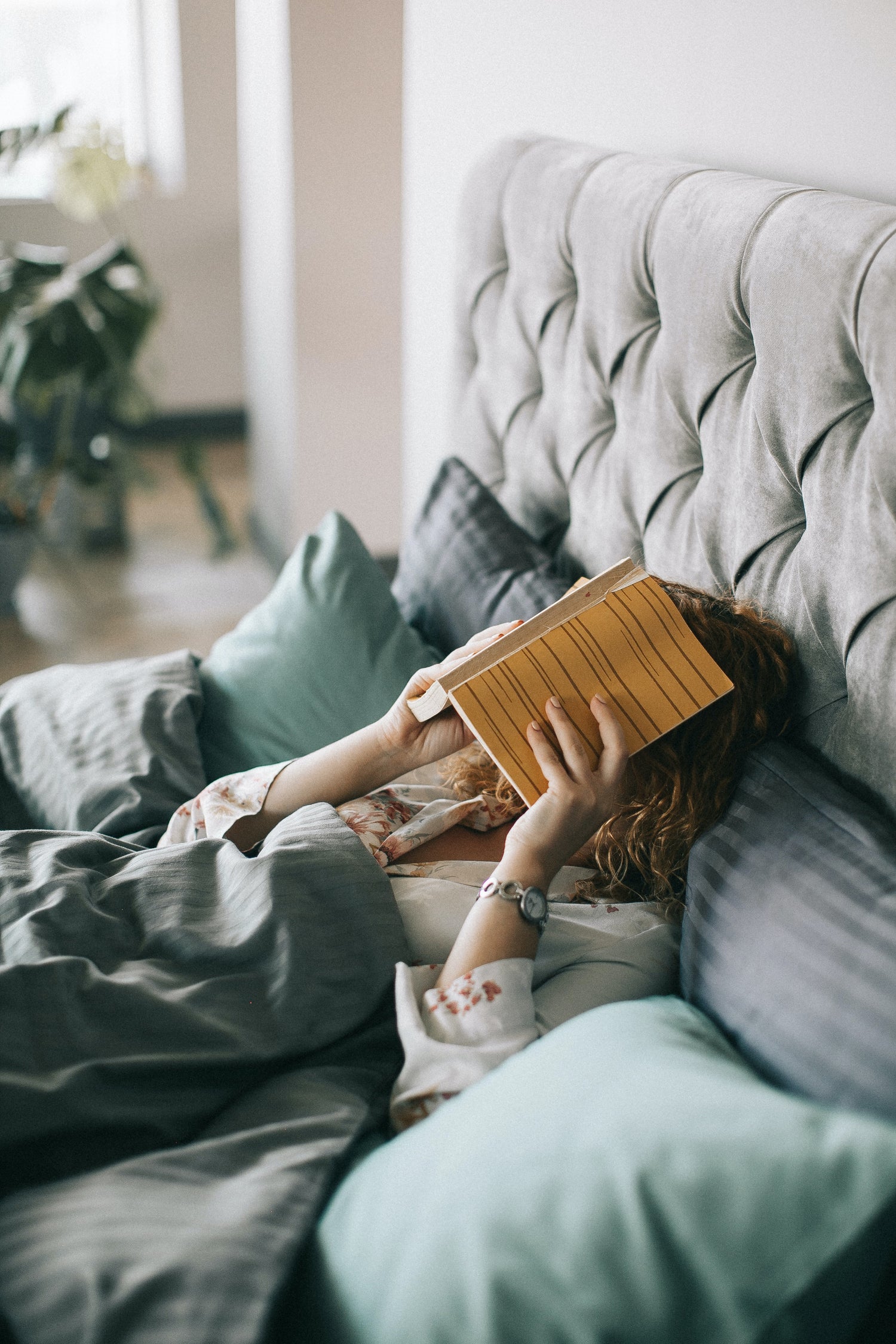 Tessie clothing. fabric headboard bed with woman in bed with a book over her face and surrounded by bedding