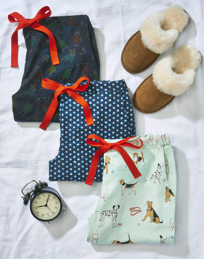 Tessie Pyjama Trousers. 3 printed pyjama sets folded onto a white waffle bed cover with sheepskin slipper mules and an alarm clock. The Rascal dog print pyjama trousers and dotty print pyjama trousers are made using Liberty Fabric and are 100% cotton. The Poppy dog print pyjama trousers made form 100% Tencel are Tessie's own design featuring Airedale Terriers, Dalmatians, Beagles and Westies on a soft green background. Tencel Pyjamas. Cotton Pyjamas